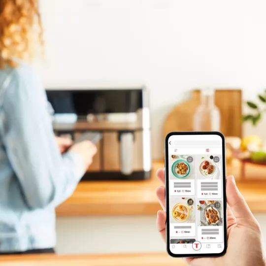 Person using Tefal Dual Easy Fry & Grill air fryer (8.3L, 2700W) in a kitchen while following recipes on a smartphone app.